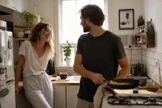 Couple laughing while making breakfast in a small sunlit kitchen